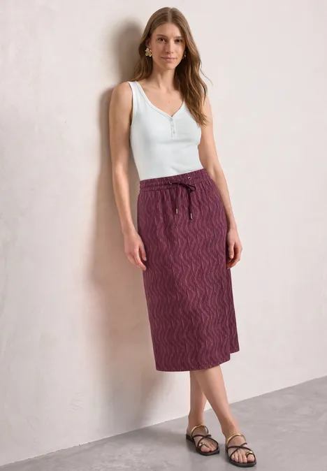 Midirock mit Elastikbund und Print blush berry Frau trägt weißes Top, violette gemusterte Midirock und schwarze Sandalen, vor hellem Hintergrund.