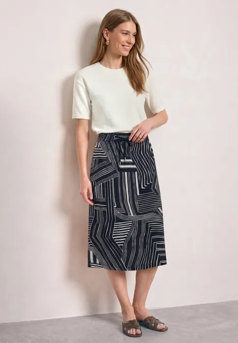Midirock mit Elastikbund und Print darkest blue Frau vor hellem Hintergrund mit weißem T-Shirt, schwarzem Rock mit geometrischem Muster und schwarzen Sandalen.
