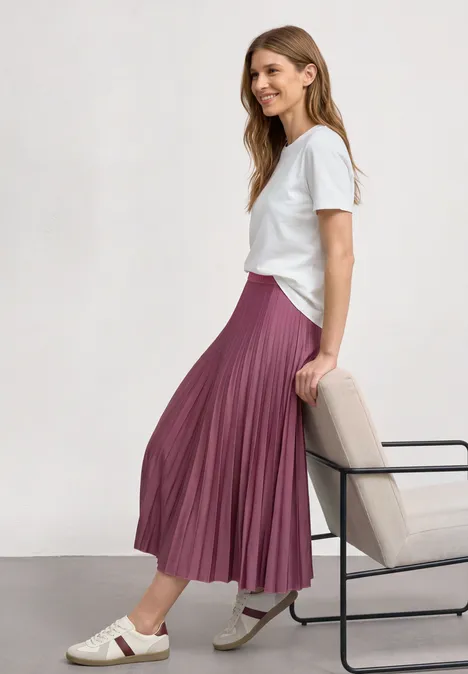 Jupe midi à structure plissée dark clouded blush Une femme porte un t-shirt blanc et une jupe plissée rose, assise de profil sur une chaise.