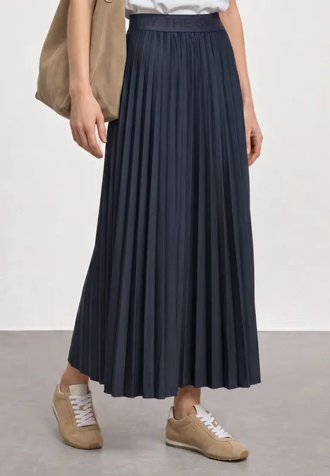 Persoon draagt een donkerblauwe geplisseerde midirok, witte blouse, beige sneakers en een schoudertas.
