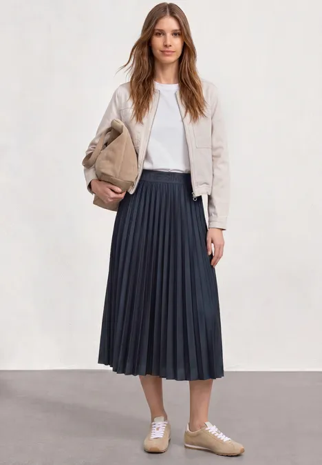 Vrouw met beige suède jack, wit shirt, midirok met plooien in donkerblauw, beige sneakers en clutch.