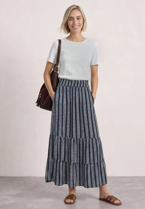 Femme en t-shirt blanc, jupe longue rayée bleu et blanc, sandales brunes et sac à l’épaule sur fond clair.