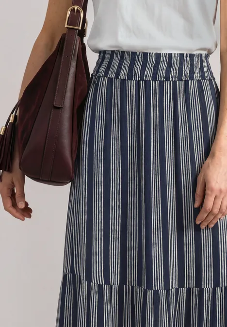 Femme en haut blanc et jupe longue rayée bleu et blanc, avec un sac marron à l’épaule.