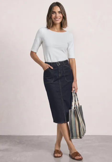Frau in weißem Oberteil, dunklem Jeans-Midirock und Sandalen hält gestreifte Tasche.