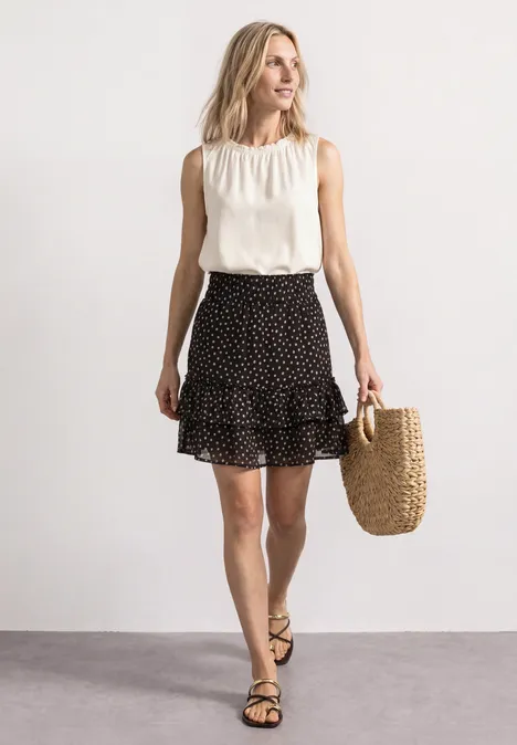 Vrouw in mouwloze witte blouse en zwarte, gestippelde rok met rieten tas en sandalen.