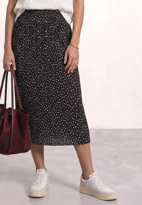 Midirock aus Mesh in Plisséestruktur black coffee Frau in weißem Top, gepunktetem schwarzen Midirock, weißen Sneakern und roter Veloursledertasche.