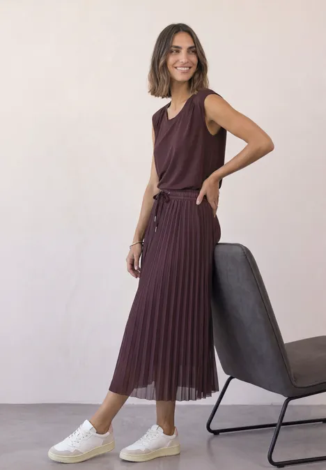 Midirock aus Mesh in Plisséestruktur rustic rouge Eine Frau in bordeauxrotem ärmellosen Oberteil und Plisseerock, weiße Sneaker, steht neben einem Stuhl.