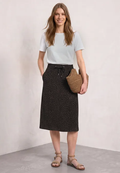 Femme en t-shirt blanc, jupe noire midi à motifs discrets, sandales et sac en osier sur fond clair.