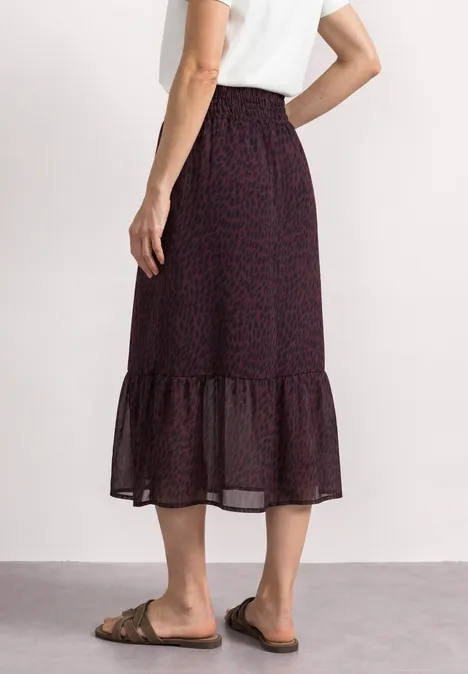 Een vrouw draagt een wit T-shirt en een donker, motief midi-rok met volant en platte sandalen.