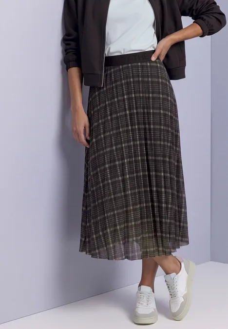 Vrouw in geruite midirok en witte sneakers, hand in heup.