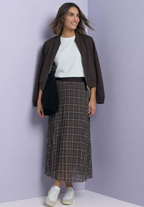 Vrouw in een bruine jas, wit topje, geruite lange rok en witte sneakers tegen een lila achtergrond.