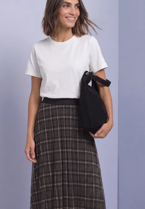 Een glimlachende vrouw voor een lila achtergrond draagt een wit T-shirt, een donker geruite rok en houdt een zwarte tas vast.