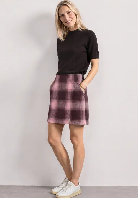 Frau in schwarzem Shirt, rosa-kariertem Minirock und weißen Sneakern vor heller Wand.