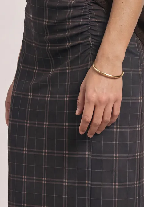 Vrouw in zwarte geruite jurk met plooidetail, arm met gouden armband zichtbaar.