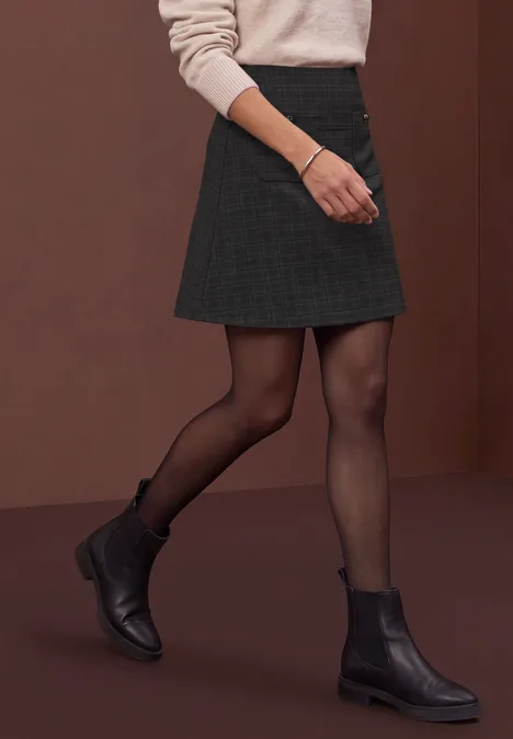 Eine Person trägt einen kurzen Rock, schwarze Strumpfhose und schwarze Stiefel vor braunem Hintergrund.