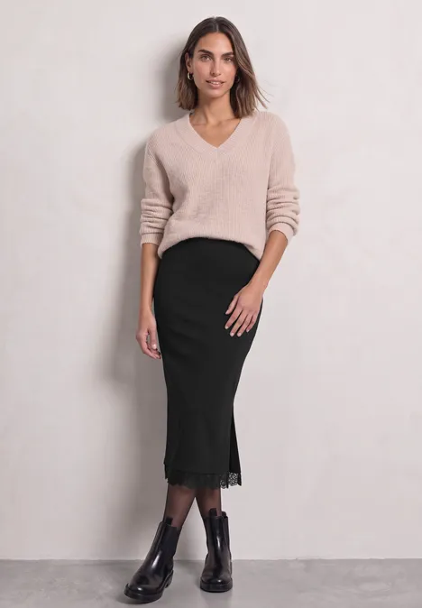 Vrouw in beige V-hals trui, zwarte midi rok met kant en enkellaarsjes voor een lichte achtergrond.