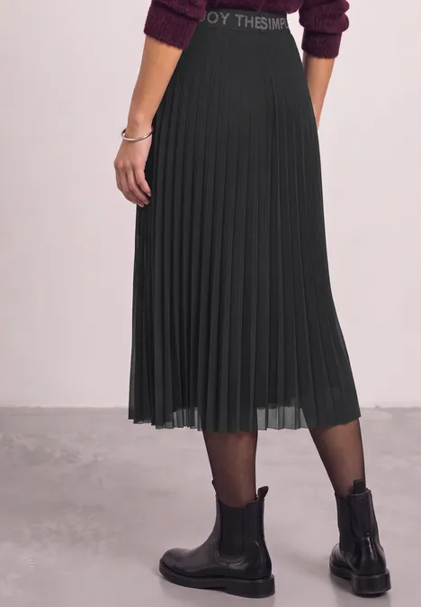 Zijaanzicht van een persoon in een zwarte, geplooide midirok met zwarte panty's en enkellaarsjes.