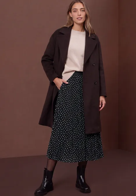 Frau mit dunkelbraunem Mantel, hellem Pullover, schwarzem gepunktetem Midirock und schwarzen Stiefeln vor braunem Hintergrund.