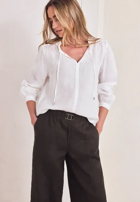 Vrouw in witte blouse en zwarte, wijde broek met riem tegen neutrale achtergrond.