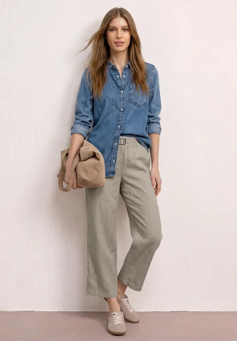 Vrouw in denimblouse, wijde broek, comfortabele schoenen, met een tas en een hoed in de hand.