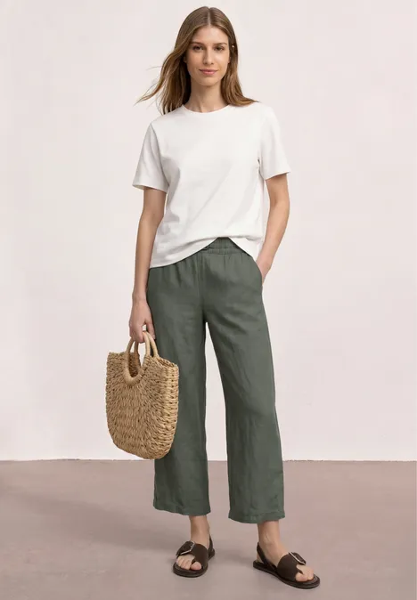 Vrouw in wit t-shirt, groengrijze linnen broek, zwarte sandalen, met gevlochten rieten tas.