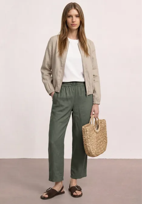 Vrouw met beige jas, wit T-shirt, groene broek, zwarte sandalen en gevlochten rieten tas.