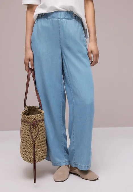 Vrouw in lichtblauwe, wijde jeans, beige loafers, wit shirt en een gevlochten stro-tas.