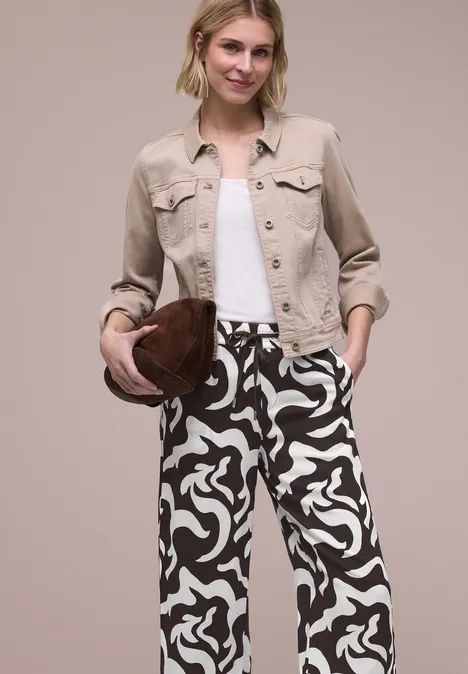 Person in gemusterter schwarz-weißer Hose, weißem Top, beigem Blazer, hält braunen Hut.