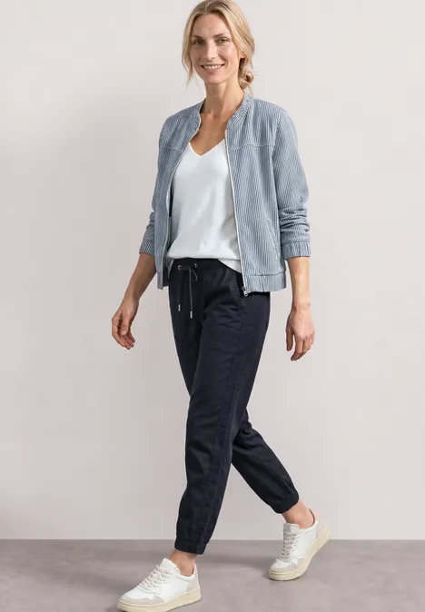 Frau in sportivem Outfit: helle Bomberjacke, weißes T-Shirt, dunkle Jogginghose und weiße Sneaker.