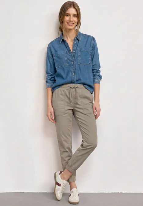 Vrouw in jeansoverhemd, beige chino en witte sneakers tegen lichte achtergrond.