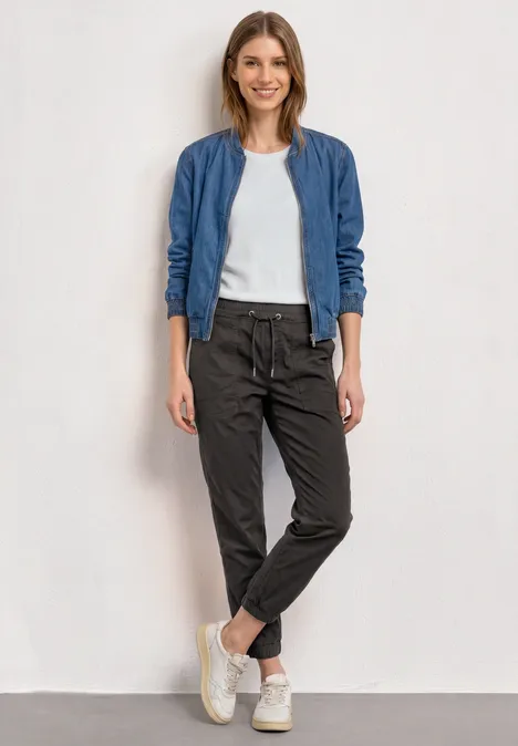 Vrouw in casual outfit met blauw jasje, wit T-shirt, zwarte broek en witte sneakers tegen neutrale achtergrond.