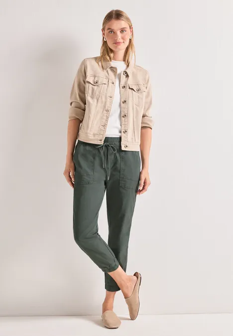 Vrouw voor neutrale achtergrond, draagt een lichtbeige jas, wit t-shirt, groene joggingbroek en slippers.