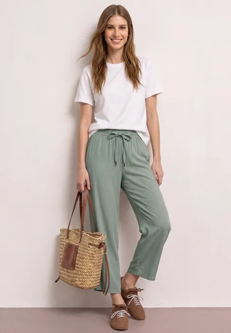 Frau lehnt an Wand, trägt weiße Bluse, grüne Hose, Basttasche und braune Schuhe.