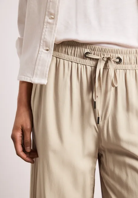 Persoon draagt beige broek met trekkoord en wit bovenstuk, hand langs het lichaam, voor een lichte achtergrond.