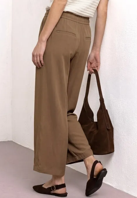 Vrouw van achteren in losse, bruine broek, witte top, zwarte sandalen en een bruine schoudertas.