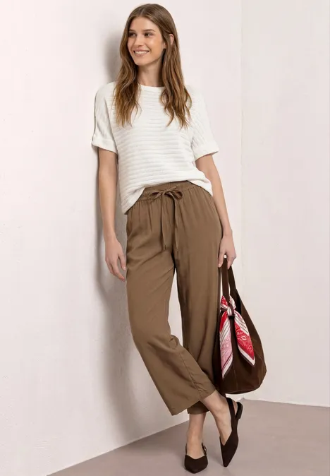 Vrouw in wit kort mouwshirt en bruine broek, met schoudertas en roze sjaal.