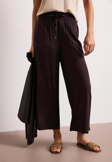 Losse, zwarte broek met trekkoord, gecombineerd met bruine sandalen en een bijpassend jasje in de hand.