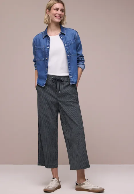 Vrouw draagt denimoverhemd, wit t-shirt en wijde grijze broek voor beige achtergrond.