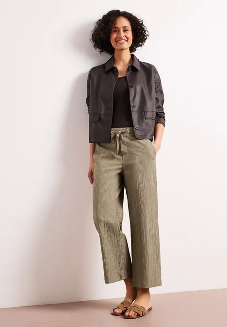 Vrouw met krullend haar draagt een zwarte trui, donker jasje, olijfgroene wijde broek en sandalen voor een lichte achtergrond.