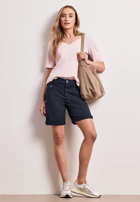Persoon in roze T-shirt, donkerblauwe bermudashorts en witte sneakers met canvas tas voor een witte muur.