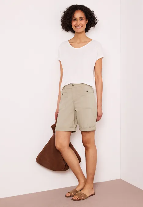 Persoon met wit t-shirt, beige shorts, bruine tas en sandalen tegen een lichte achtergrond.