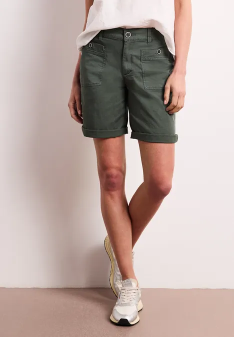 Persoon met olijfgroene korte broek en witte sneakers voor een lichte achtergrond.