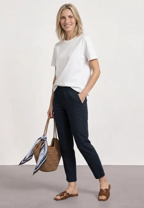 Slim Leg Chino mit Elastikbund shadowed navy Frau in weißem T-Shirt, dunklen Hosen, geflochtenen Sandalen und Strohtasche mit Tuch.