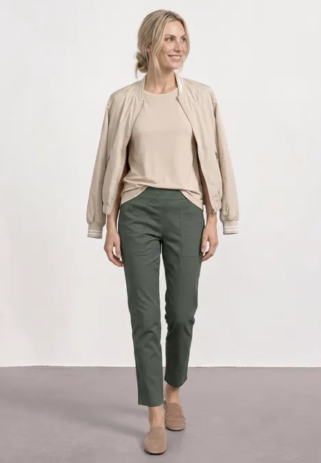 Eine Frau trägt eine beige Jacke, ein beiges Shirt, dunkelgrüne Hose und beige Schuhe vor neutralem Hintergrund.