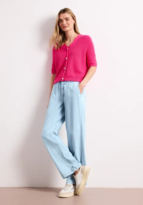 Dame in pinkem Cardigan, hellblauer Marlenehose und weißen Sneakers, steht entspannt vor hellem Hintergrund.