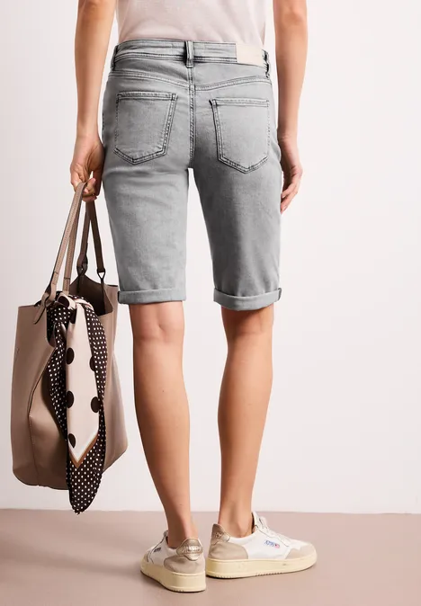 Achteraanzicht van een persoon in lichtgrijze denim bermudashorts, met beige sneakers en een tas met gestipte sjaal.