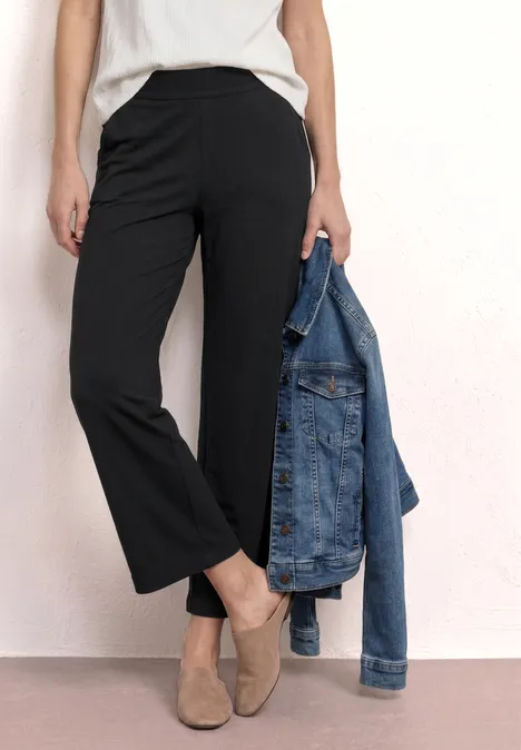 Unterkörper einer Person mit schwarzen weiten Hosen, beigen Schuhen und einer Jeansjacke in der Hand.