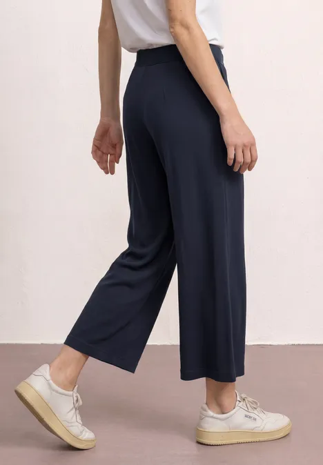 Eine Person trägt weite, knöchellange, dunkelblaue Hosen und weiße Sneaker.