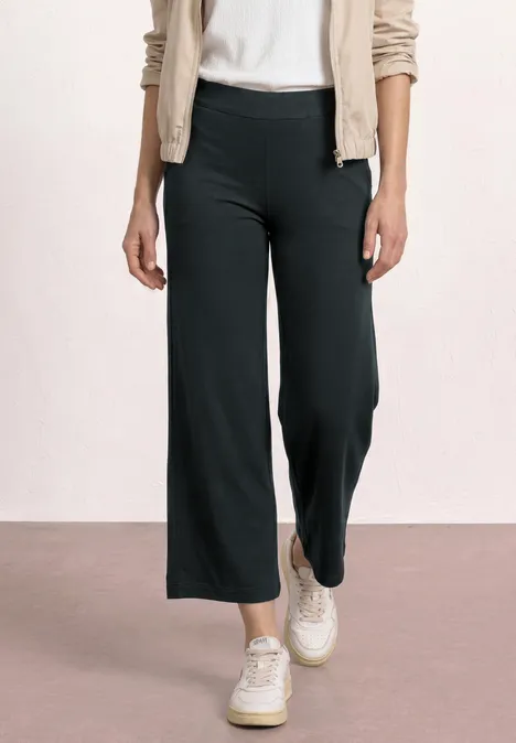 7/8 Wide Leg Hose im Loose Fit pine grove green Frau trägt schwarze, weite 7/8-Hose mit weißen Sneakers und beiger Strickjacke