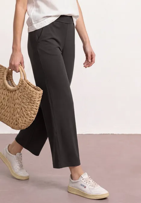 Schlanke 7/8-Hose in Schwarz, weiße Sneakers und Basttasche auf hellem Boden